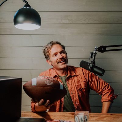 maple Man holds football in his podcast room in front of maple wood wall.